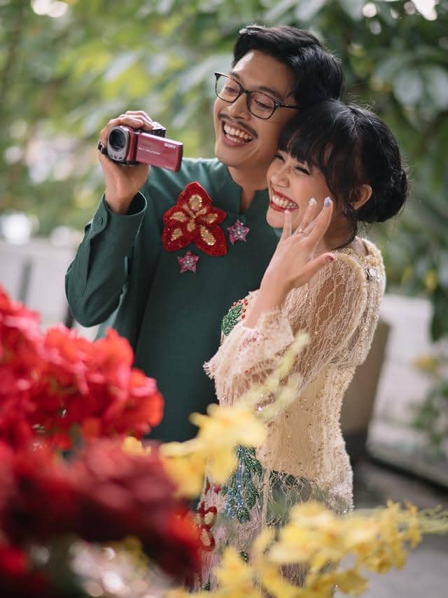Sepasang kekasih tersenyum bahagia saat merekam momen lamaran, dikelilingi bunga berwarna merah dan kuning yang cerah.