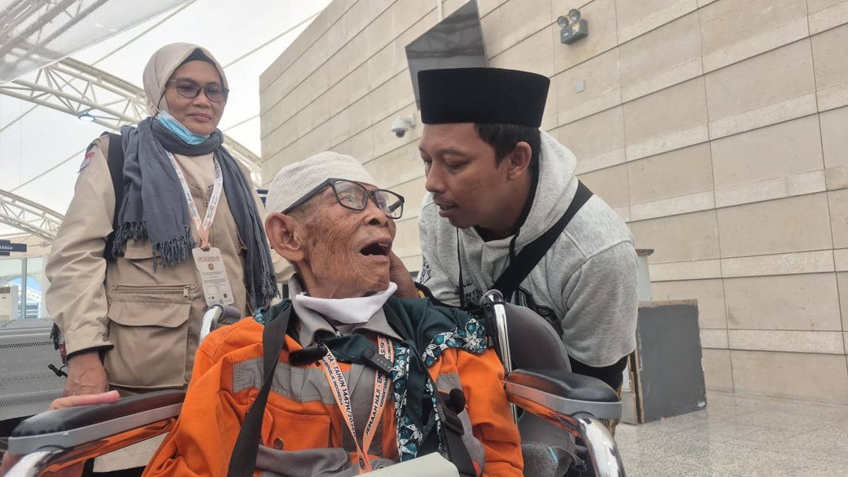 Seorang jemaah lanjut usia duduk di kursi roda berbicara dengan pendampingnya, sementara seorang perempuan berdiri di samping mereka di area bandara.