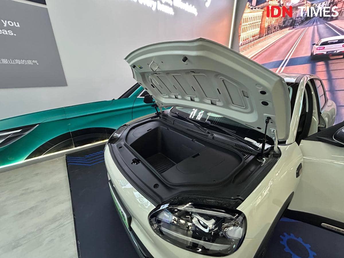 Bagasi depan mobil listrik Chery Q berwarna putih terbuka di area pameran dengan latar belakang mobil lain dan layar besar.