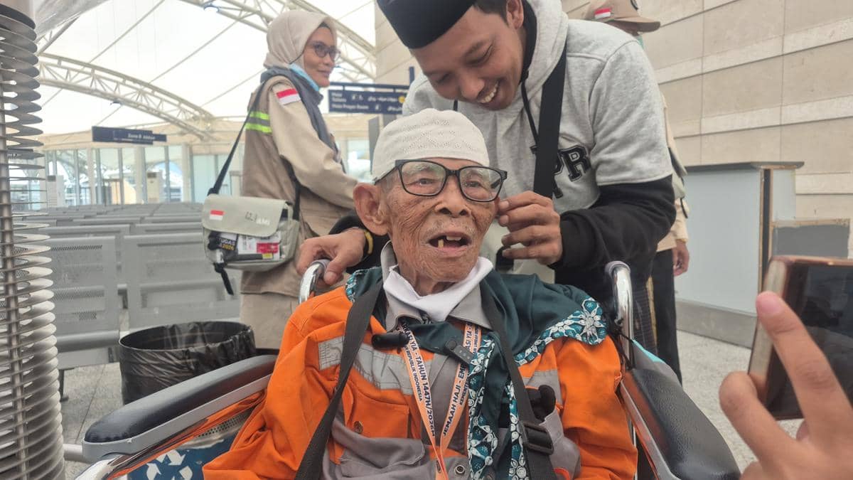 Seorang jemaah lanjut usia duduk di kursi roda mengenakan seragam oranye haji, tersenyum saat didampingi oleh seorang pria muda di Bandara Madinah.