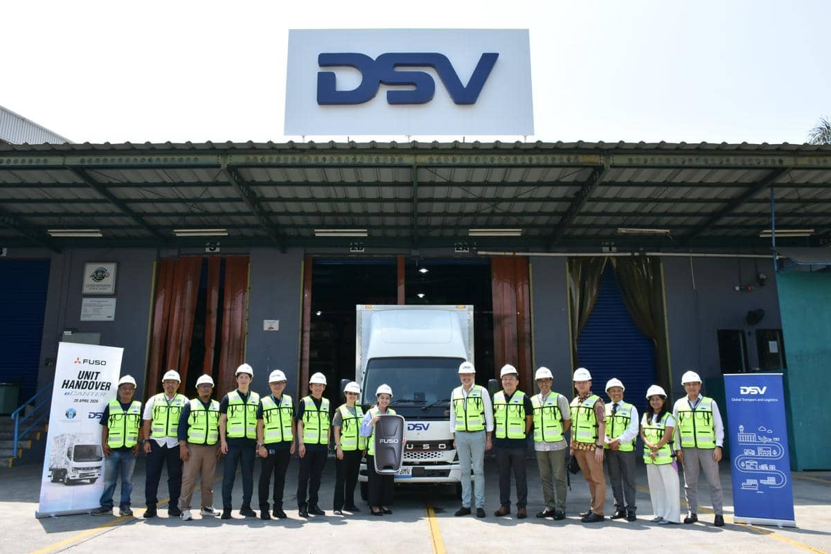 Sejumlah orang mengenakan rompi keselamatan dan helm berdiri di depan truk Fuso di area gudang dengan logo DSV di atasnya.