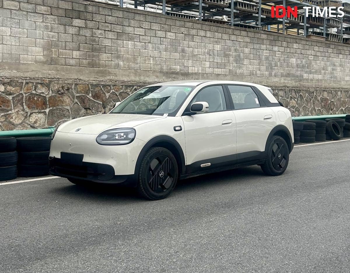 Mobil SUV berwarna putih dengan desain membulat di berbagai sisi sedang diparkir di area jalan dekat dinding batu dan tumpukan ban.