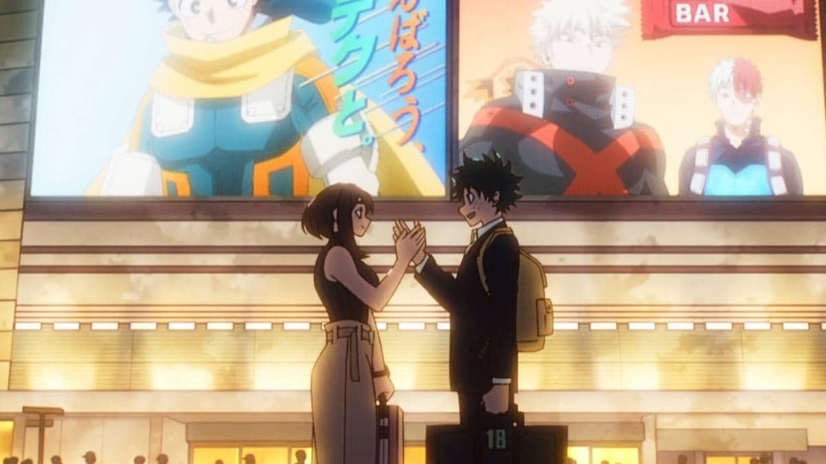 Midoriya dan Uraraka berdiri saling berhadapan di depan layar besar yang menampilkan pahlawan dari serial My Hero Academia.