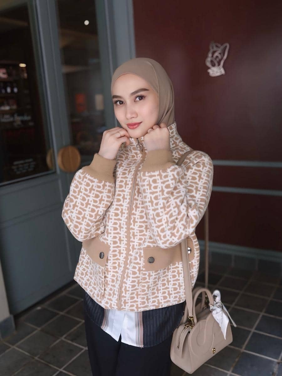 Melody Laksani berhijab mengenakan jaket motif krem dan membawa tas tangan berwarna senada, berpose di depan pintu kaca dan dinding merah.