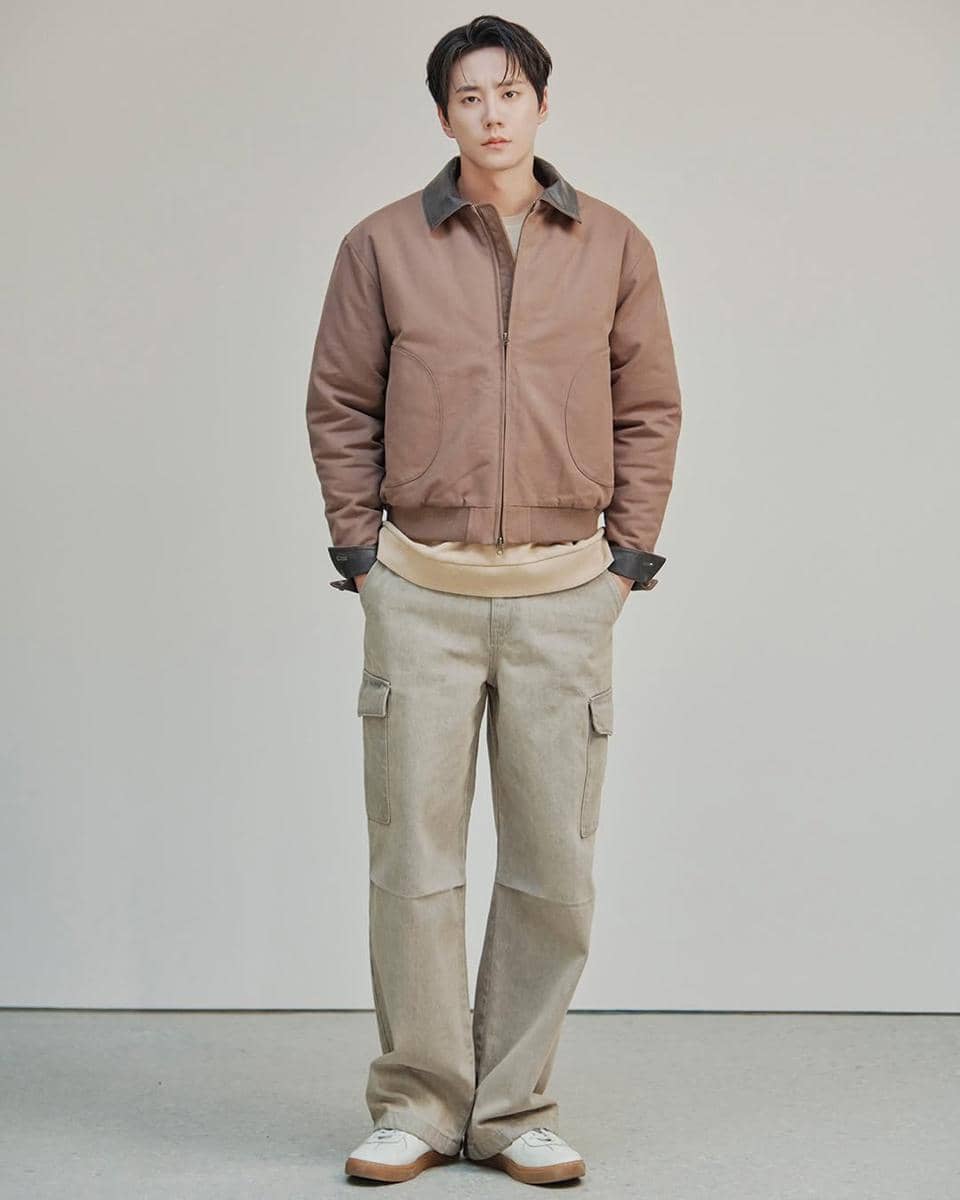 Seorang pria berdiri mengenakan jaket cokelat muda, celana panjang krem dengan kantong samping, dan sepatu putih di ruangan minimalis.