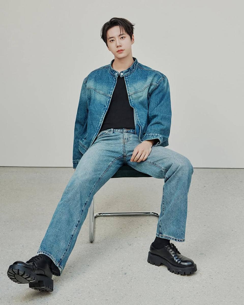 Seorang pria duduk di kursi mengenakan jaket denim biru, kaus hitam, celana jeans, dan sepatu hitam tebal di ruangan minimalis.