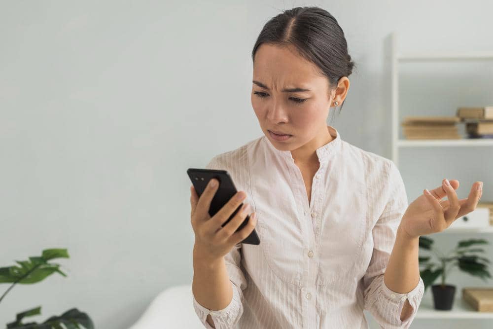 Seorang wanita mengenakan kemeja putih memegang ponsel dengan ekspresi bingung di ruangan dengan latar belakang rak dan tanaman.