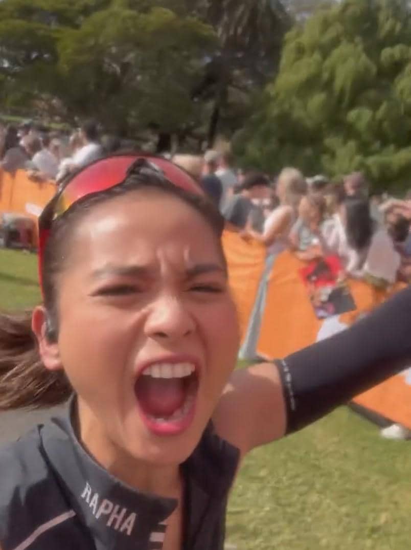 Seorang pelari perempuan berteriak penuh semangat di area garis finis Sydney Half Marathon dengan latar penonton di belakangnya.