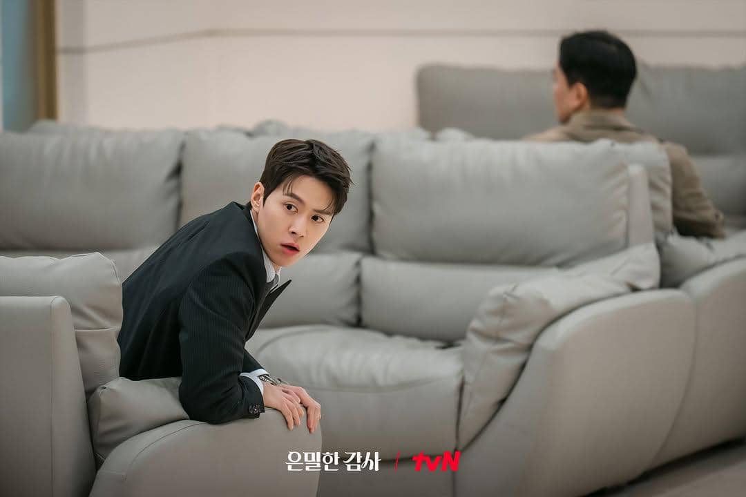 Seorang pria muda mengenakan setelan jas hitam duduk di sofa abu-abu dengan ekspresi terkejut di adegan drama Filling for Love.