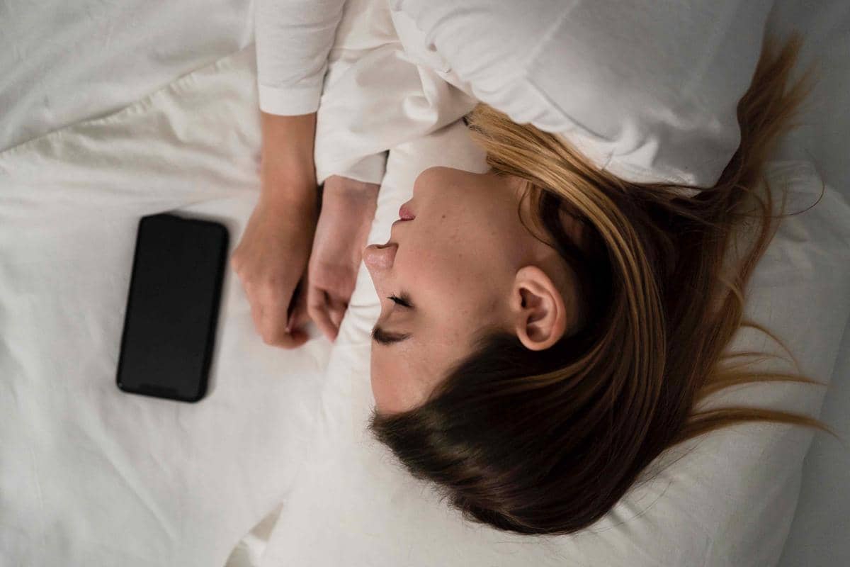 Seorang wanita berbaring di tempat tidur dengan mata terpejam, mengenakan pakaian putih, dan ponsel tergeletak di sampingnya.