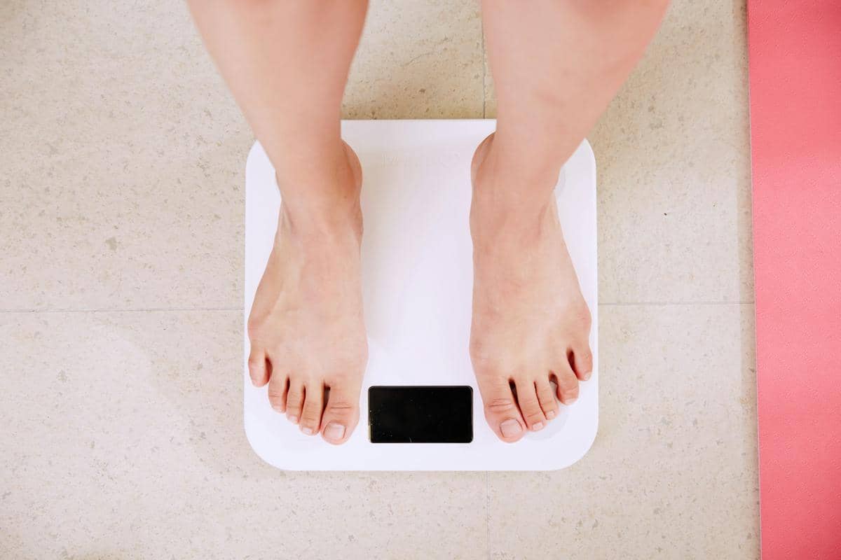 Seseorang berdiri di atas timbangan digital berwarna putih di lantai, memperlihatkan kedua kaki tanpa alas kaki dari sudut atas.