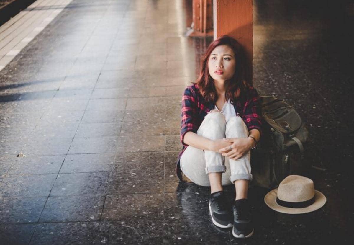 Seorang wanita duduk di lantai dekat tiang dengan ekspresi termenung, di sampingnya terdapat ransel dan topi berwarna krem.