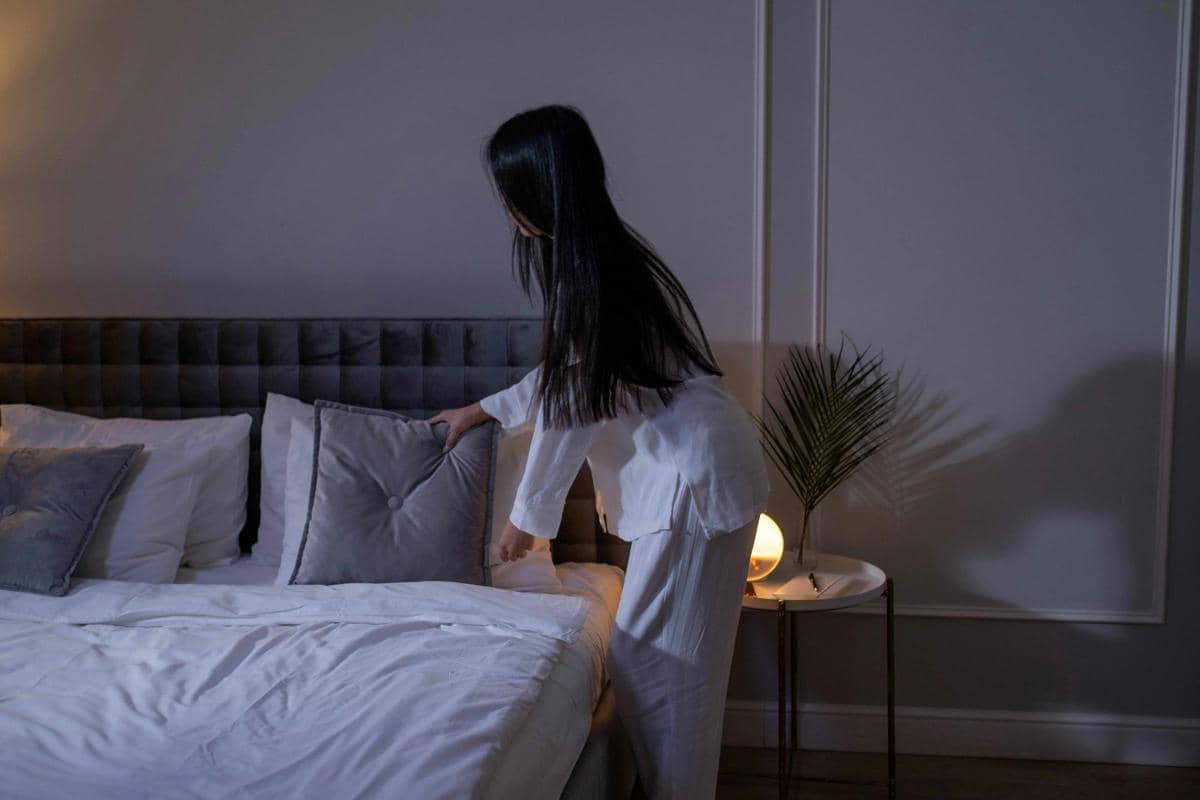 Seorang perempuan berpakaian piyama putih sedang merapikan bantal di atas tempat tidur di kamar bernuansa lembut dengan pencahayaan hangat.