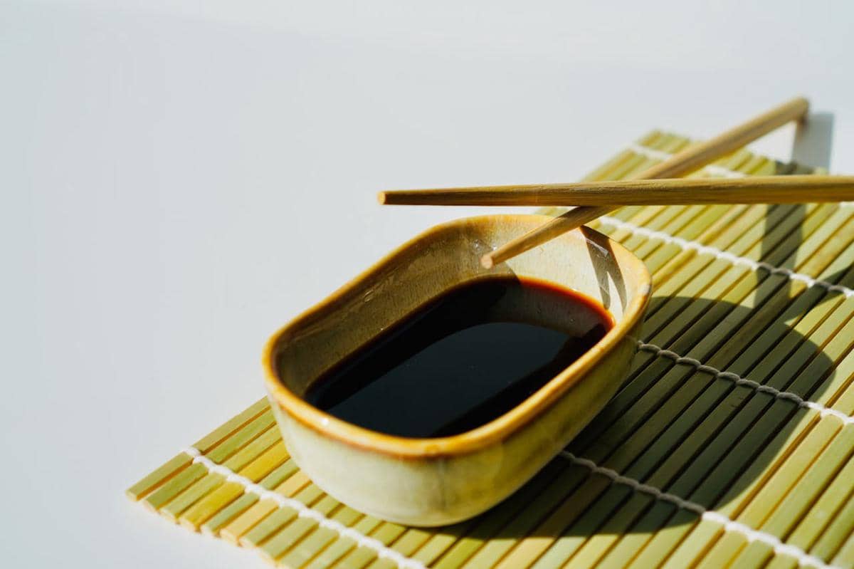 Semangkuk kecil kecap asin berwarna gelap dengan sepasang sumpit kayu di atas alas bambu berwarna hijau muda.