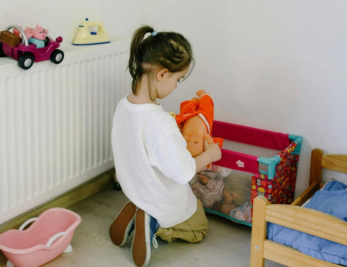 Seorang anak kecil berlutut di lantai sambil memegang boneka bayi berwarna oranye di dekat boks mainan di kamar bermain.