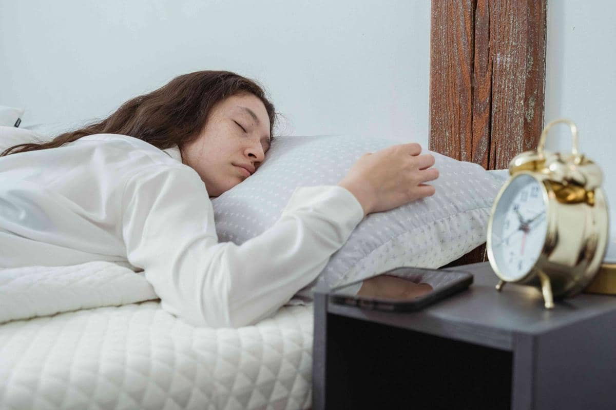 Seorang perempuan tidur di tempat tidur dengan piyama putih, ponsel diletakkan di meja samping bersama jam weker berwarna emas.