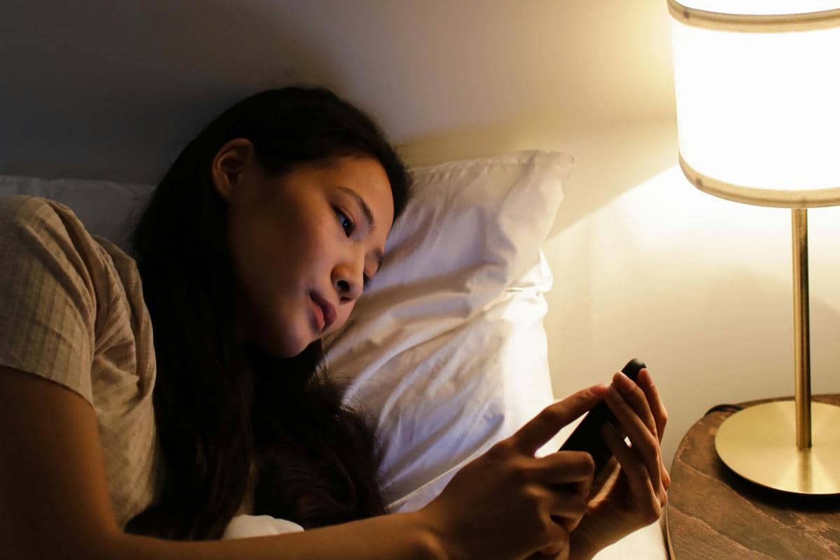 Seorang perempuan berbaring di tempat tidur sambil menggunakan ponsel di bawah cahaya lampu meja di kamar yang redup.