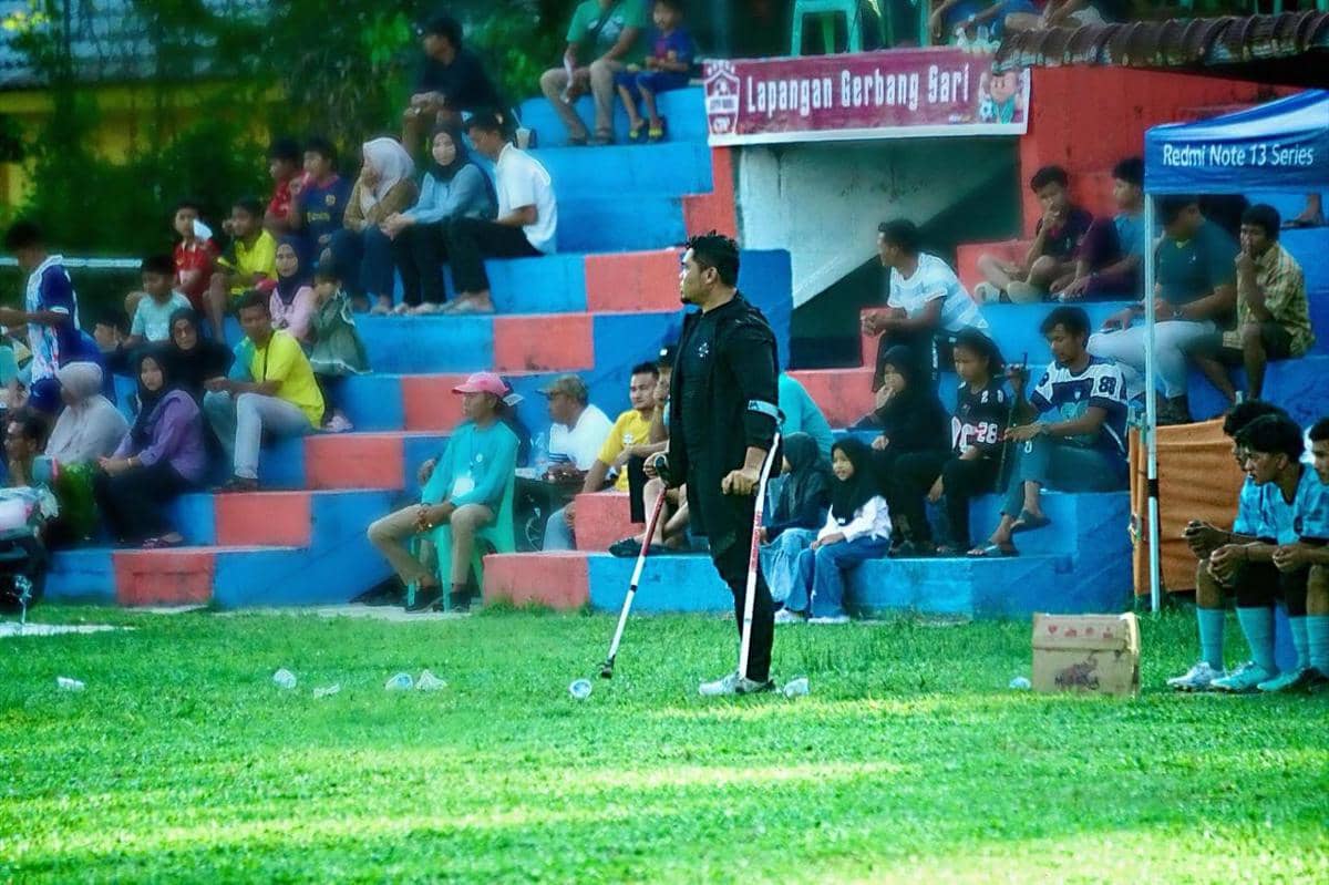 Seorang pelatih berdiri di pinggir lapangan dengan tongkat, mengarahkan tim sepak bola muda di depan penonton di tribun stadion.