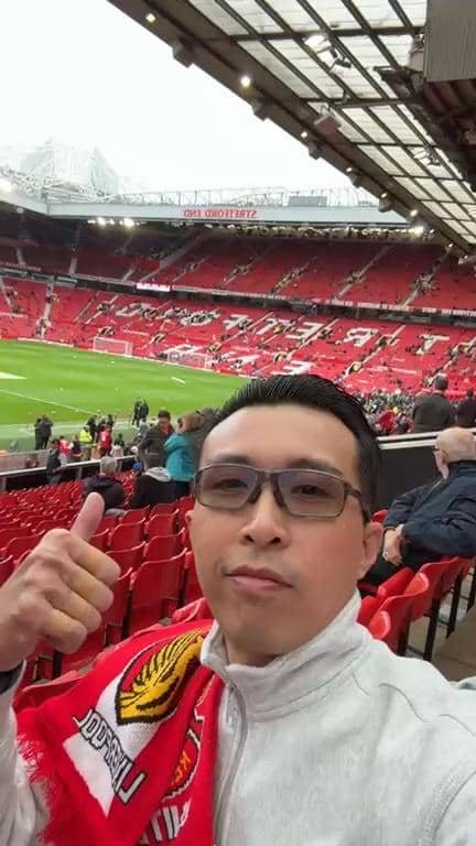 dr. Tirta berpose dengan jempol terangkat di tribun stadion Old Trafford yang dipenuhi penonton sebelum pertandingan dimulai.