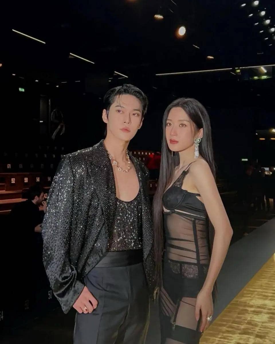 Seorang pria dan wanita berpose di panggung fashion show dengan pencahayaan mewah, mengenakan busana hitam elegan dan berkilau.