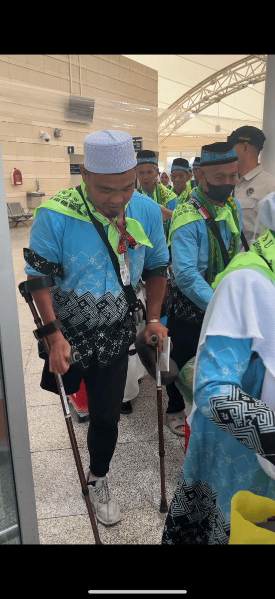 Seorang jemaah haji mengenakan pakaian seragam biru bermotif hitam putih dengan syal hijau, berjalan menggunakan kruk di area keberangkatan.