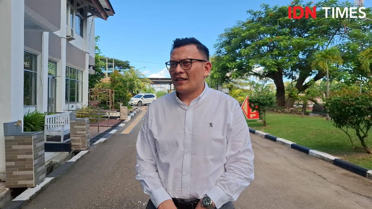 Seorang pria mengenakan kemeja putih dan kacamata berdiri di luar gedung dengan latar pepohonan hijau dan jalan berpaving.
