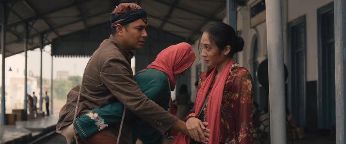 Adegan dari film Badut Gendong memperlihatkan tiga orang berpakaian tradisional di stasiun dengan ekspresi emosional.