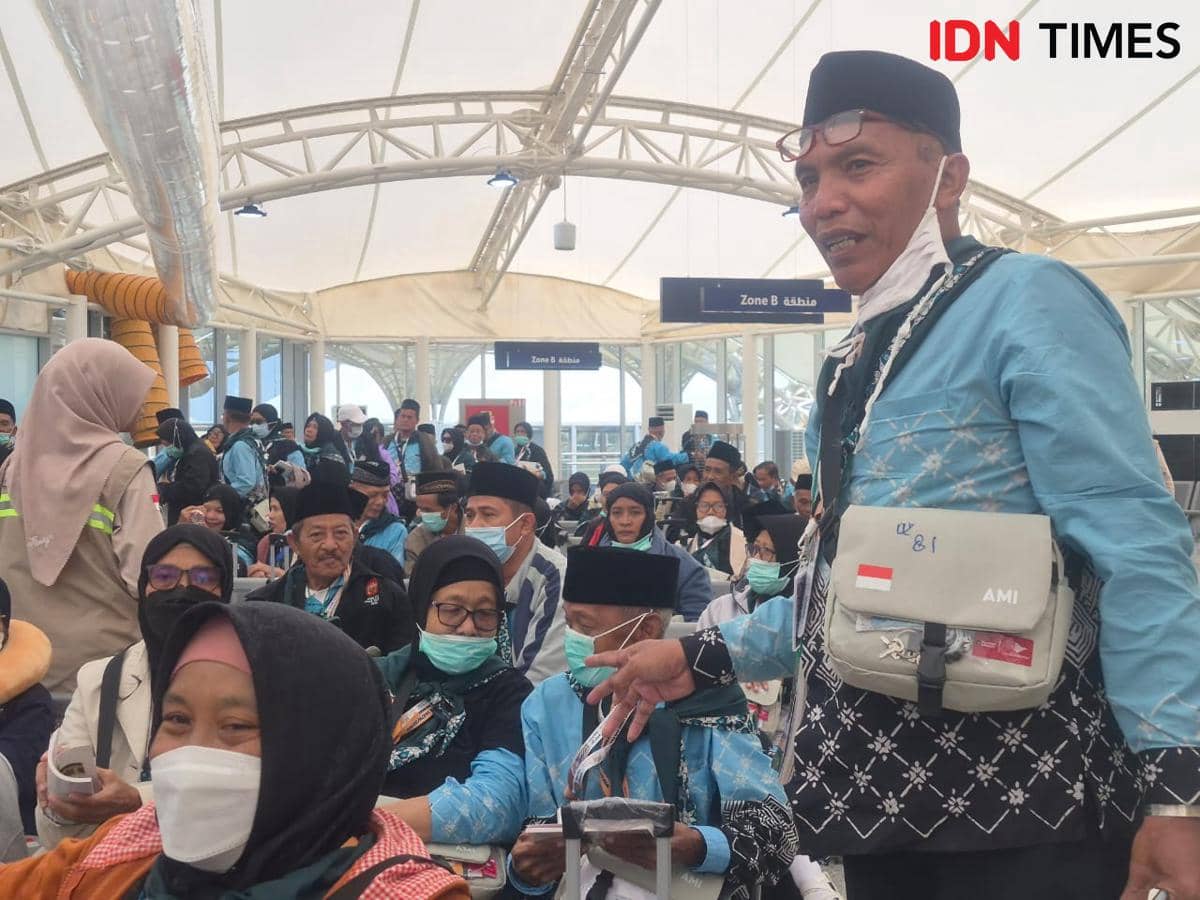 Seorang jemaah haji mengenakan seragam biru muda bersama rombongan Kloter YIA-9 menunggu di area keberangkatan dengan suasana ramai.