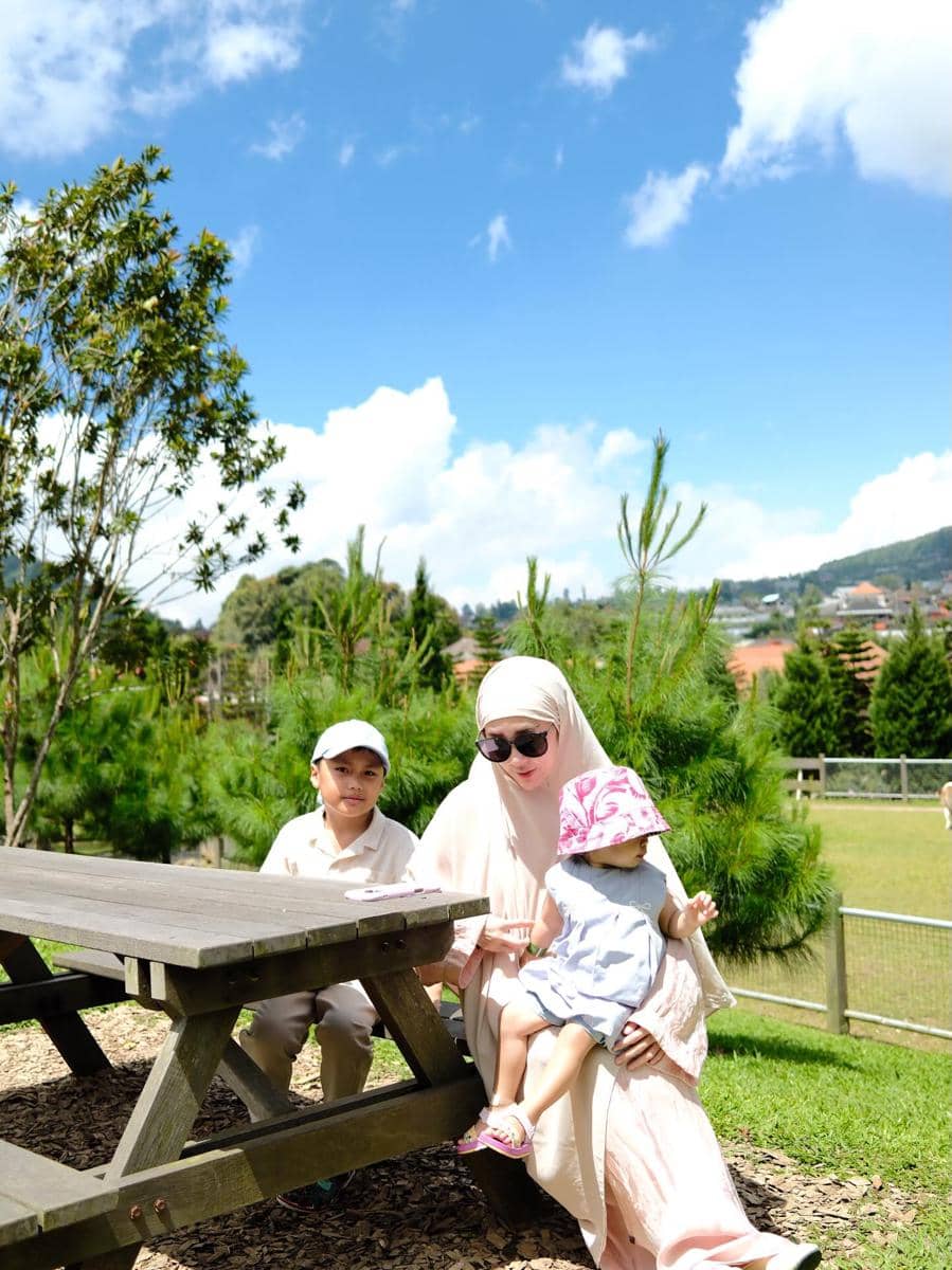 Larissa Chou duduk di bangku taman bersama dua anak kecil di bawah langit cerah dengan latar pepohonan hijau.
