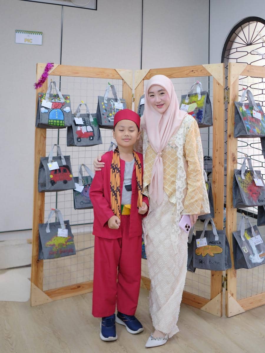 Larissa Chou berdiri bersama anak laki-lakinya yang mengenakan pakaian tradisional merah di depan pajangan tas kain hasil karya anak-anak.