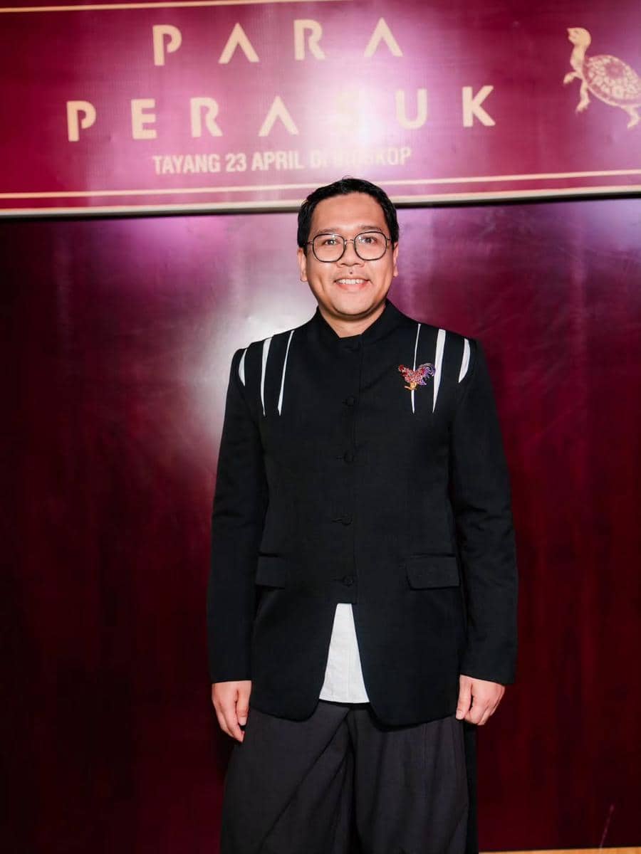 Seorang pria mengenakan jas hitam dengan detail garis putih dan bros di dada, berdiri di depan latar bertuliskan 'Para Perasuk'.