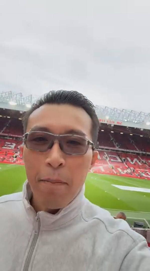 dr. Tirta mengenakan jaket abu-abu dan kacamata berfoto selfie di dalam stadion Old Trafford dengan latar tribun merah dan lapangan hijau.