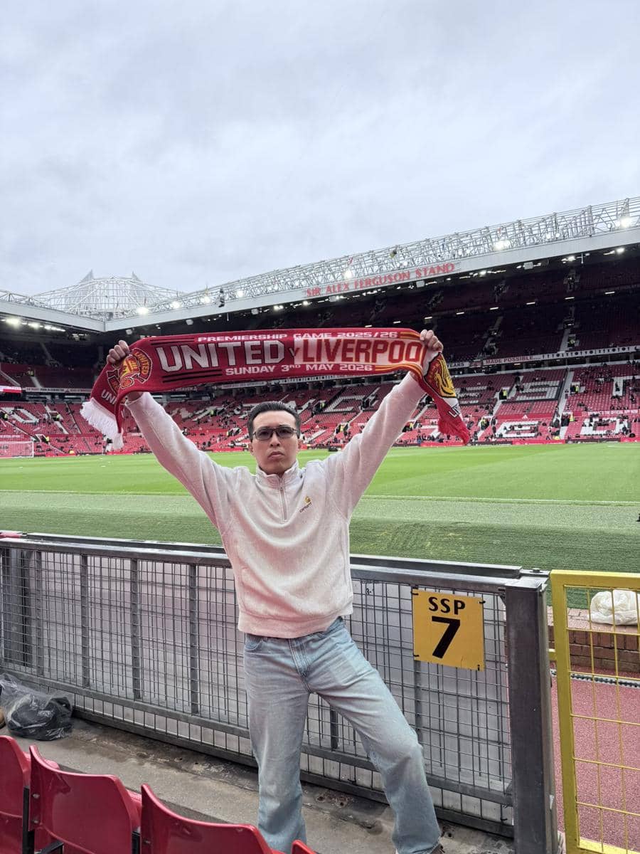  mengenakan sweter putih dan celana jeans berdiri di tribun Old Trafford sambil mengangkat syal pertandingan Manchester United vs Liverpool.