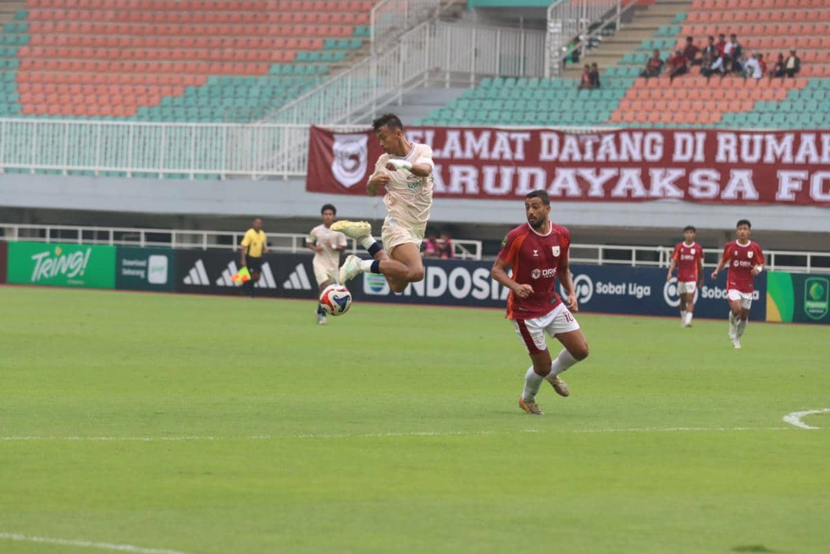 Pemain Garudayaksa FC melompat mengontrol bola saat pertandingan melawan tim berbaju merah di stadion dengan tribun kosong sebagian.