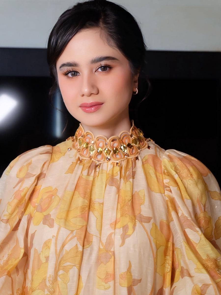 Seorang perempuan mengenakan gaun bermotif bunga warna kuning dan oranye dengan riasan lembut serta kalung besar berwarna emas.