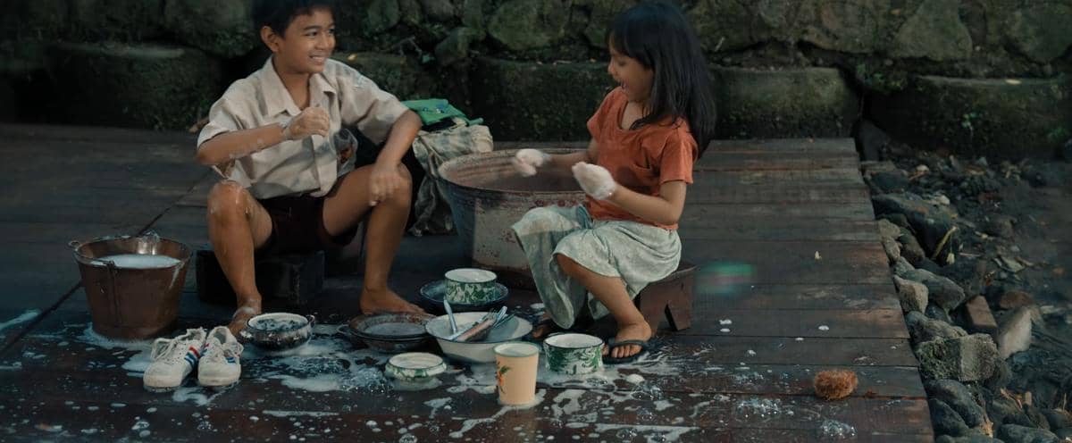 Dua anak kecil duduk di lantai kayu sambil mencuci piring dan sepatu dengan sabun, tampak tersenyum dan bermain busa bersama.
