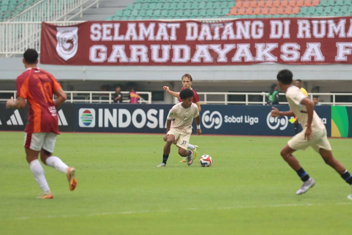 Pemain Garudayaksa FC menggiring bola saat pertandingan di stadion dengan spanduk bertuliskan sambutan untuk tim di latar belakang.
