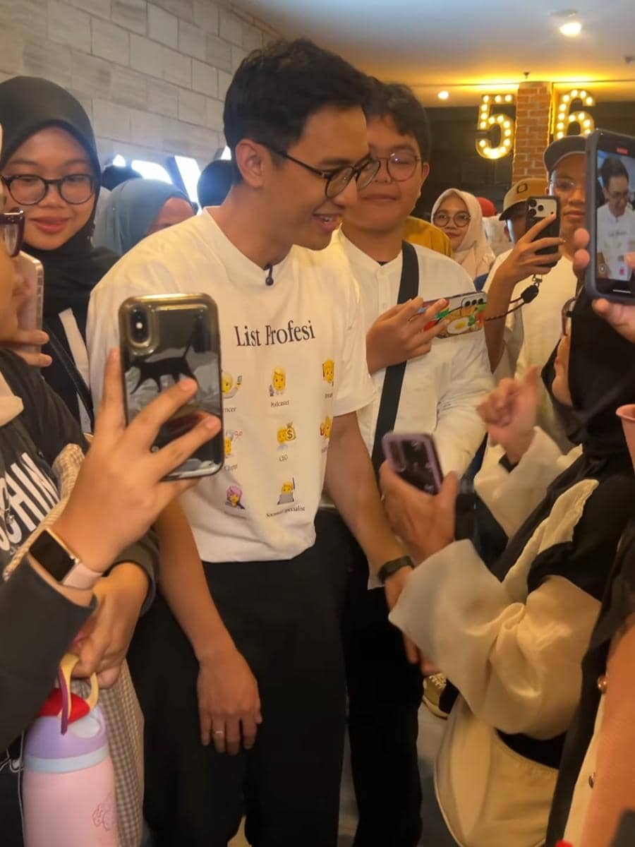 Seorang pria mengenakan kaus putih bertuliskan 'List Profesi' dikelilingi penonton yang mengambil foto di acara pemutaran film.