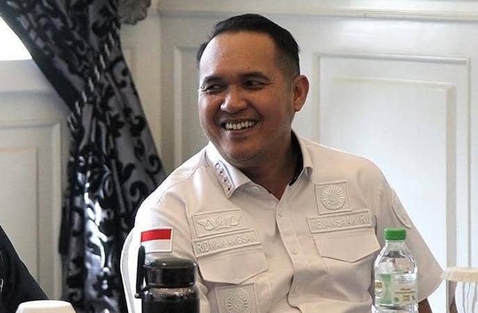 Seorang pria mengenakan kemeja putih dengan bendera Indonesia di lengan kiri sedang tersenyum saat duduk di ruangan bercat putih.