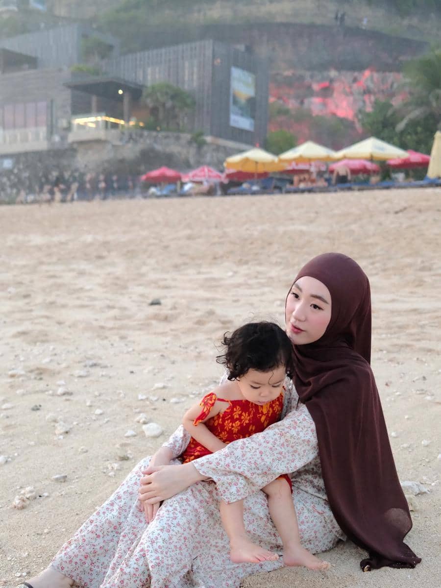Larissa Chou duduk di pasir pantai sambil memeluk anak kecil yang mengenakan pakaian merah bermotif bunga.