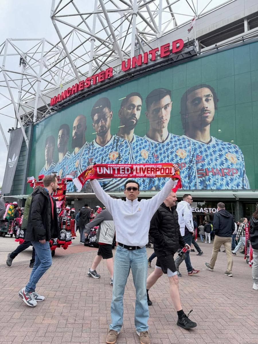  berdiri di depan Stadion Old Trafford sambil mengangkat syal bertuliskan United Liverpool, dengan latar belakang mural pemain Manchester United.