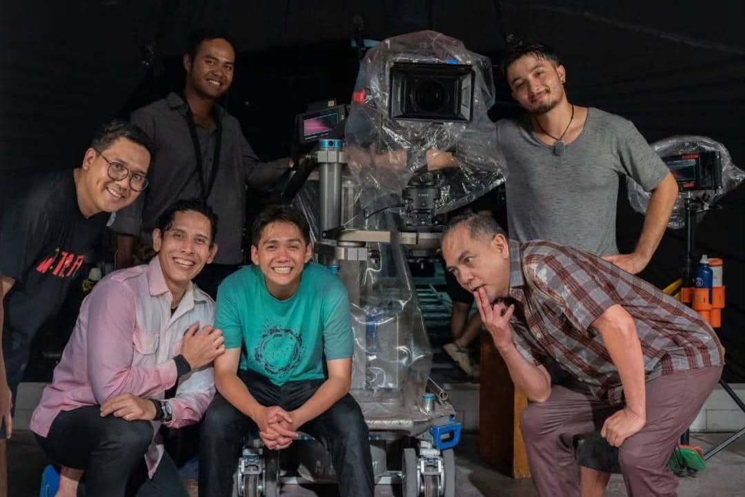 Enam kru film tersenyum di sekitar kamera film besar yang dilapisi plastik di lokasi syuting film Para Perasuk.