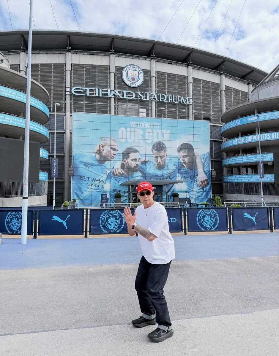 dr. Tirta berpose di depan Etihad Stadium, markas Manchester City, dengan latar mural pemain sepak bola dan langit cerah.