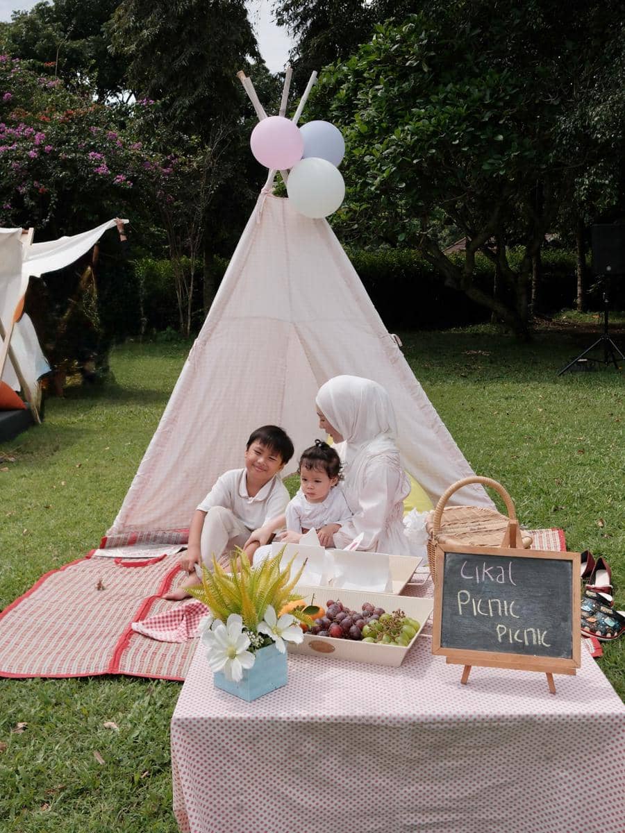 Larissa Chou duduk bersama dua anak di depan tenda kecil dengan dekorasi balon dan makanan piknik di taman hijau.