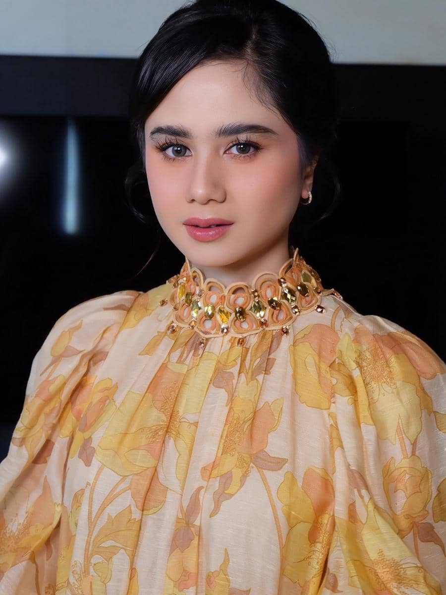 Seorang wanita mengenakan gaun bermotif bunga warna kuning dan oranye dengan riasan wajah lembut serta rambut disanggul rapi.