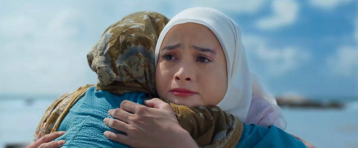 Seorang perempuan berhijab memeluk seseorang sambil meneteskan air mata di tepi laut dengan latar langit biru cerah.