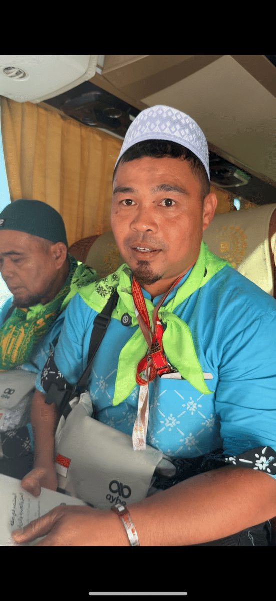 Seorang jemaah haji mengenakan pakaian biru dengan syal hijau muda dan tas selempang duduk di dalam bus bersama jemaah lain.