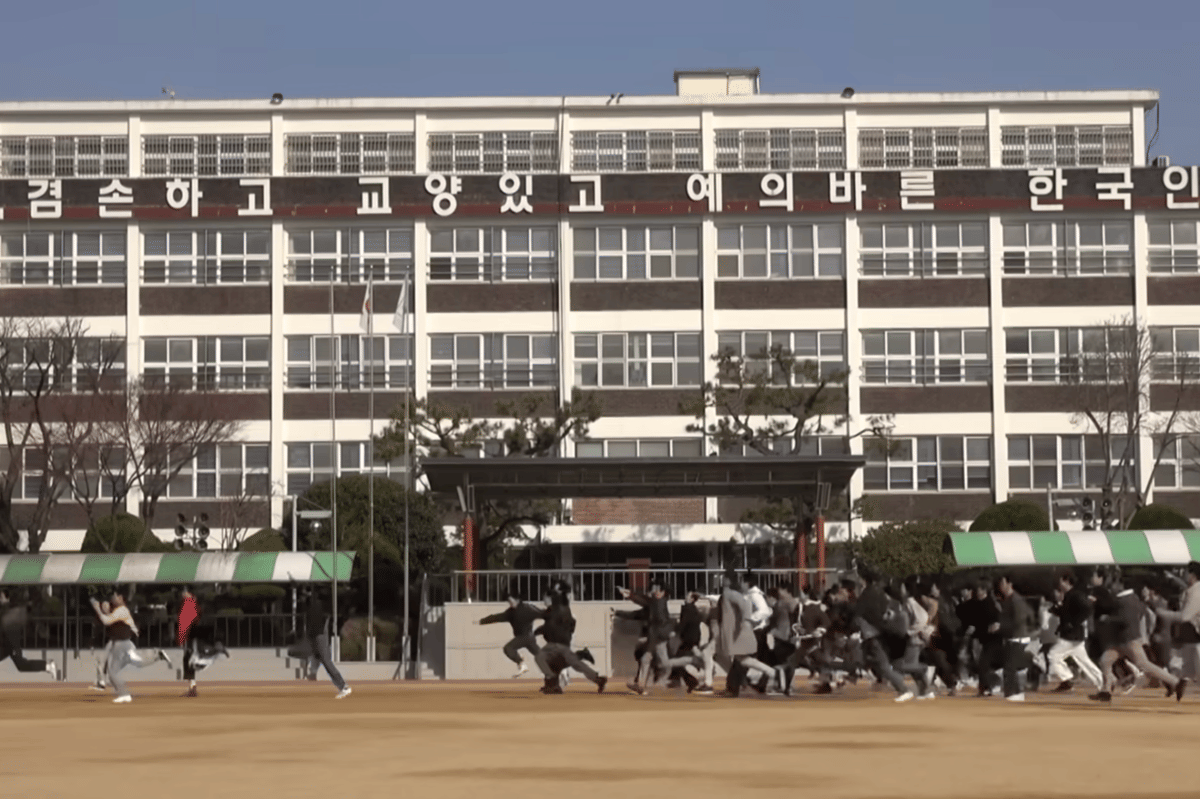 Sekelompok orang berlari di halaman sekolah besar bertingkat dengan tulisan Korea di bagian atas gedung pada siang hari yang cerah.