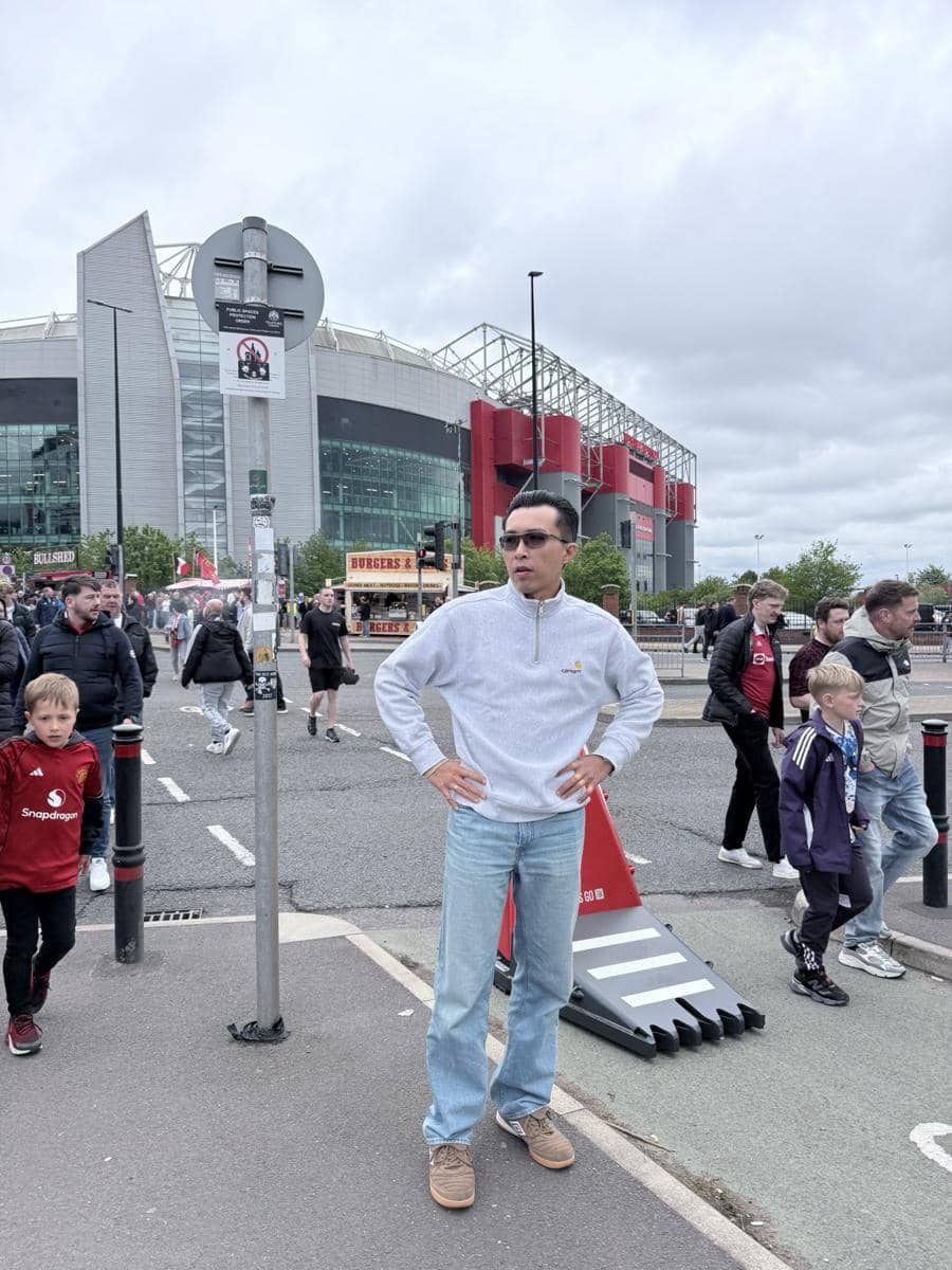 dr. Tirta mengenakan sweter abu-abu dan celana jeans berdiri di depan stadion Old Trafford dengan suasana ramai pengunjung.