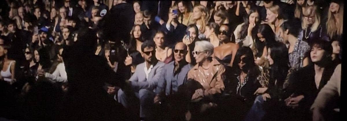 Penonton duduk di barisan depan acara fashion show dengan pencahayaan redup, menampilkan suasana ramai dan penuh perhatian.